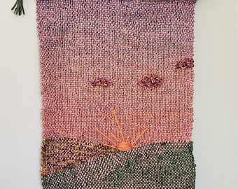May include: Handwoven wall hanging featuring a sunset scene. The tapestry has a pink and purple sky, an orange sun, and green and brown landscape. The piece is finished with a fringe and a wooden rod for hanging.