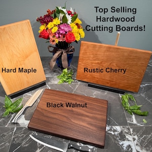 May include: Three hardwood cutting boards: Hard Maple, Rustic Cherry, and Black Walnut. The boards are displayed with a bouquet of flowers and knives. Text reads "Top Selling Hardwood Cutting Boards!"