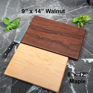 May include: A dark brown walnut cutting board measuring 9 inches by 14 inches and a light tan maple cutting board measuring 8 inches by 12 inches. A silver chef knife with a black handle and two sprigs of fresh green basil are positioned nearby.