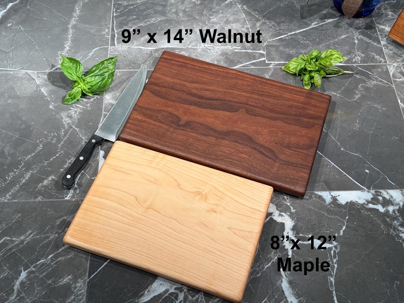 May include: A dark brown walnut cutting board measuring 9 inches by 14 inches and a light tan maple cutting board measuring 8 inches by 12 inches. A silver chef knife with a black handle and two sprigs of fresh green basil are positioned nearby.