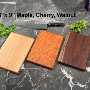 May include: Three rectangular wooden cutting boards measuring 6 inches by 9 inches, crafted from light maple, reddish cherry, and dark walnut wood. A stainless steel chef knife with a black handle rests above the boards, accompanied by fresh green basil leaves.