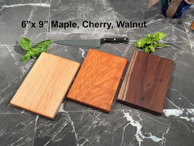 May include: Three rectangular wooden cutting boards measuring 6 inches by 9 inches, crafted from light maple, reddish cherry, and dark walnut wood. A stainless steel chef knife with a black handle rests above the boards, accompanied by fresh green basil leaves.