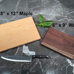 May include: A light-colored 8" x 12" maple cutting board and a dark-colored 6" x 9" walnut cutting board. A silver chef's knife with a black handle is positioned below the boards. A small sprig of green basil leaves sits between the two boards.