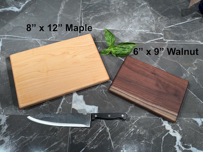 May include: A light-colored 8" x 12" maple cutting board and a dark-colored 6" x 9" walnut cutting board. A silver chef's knife with a black handle is positioned below the boards. A small sprig of green basil leaves sits between the two boards.