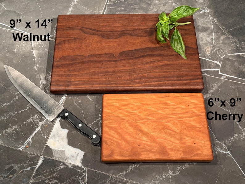 May include: A rectangular dark brown walnut cutting board measuring 9 inches by 14 inches and a smaller rectangular light orange cherry cutting board measuring 6 inches by 9 inches, accompanied by a stainless steel chef knife with a black handle.