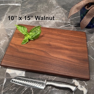 May include: A rectangular walnut cutting board measuring 10 inches by 15 inches. The dark brown wood board features a natural grain pattern and is topped with a small sprig of fresh green basil leaves. A stainless steel santoku knife is placed below.