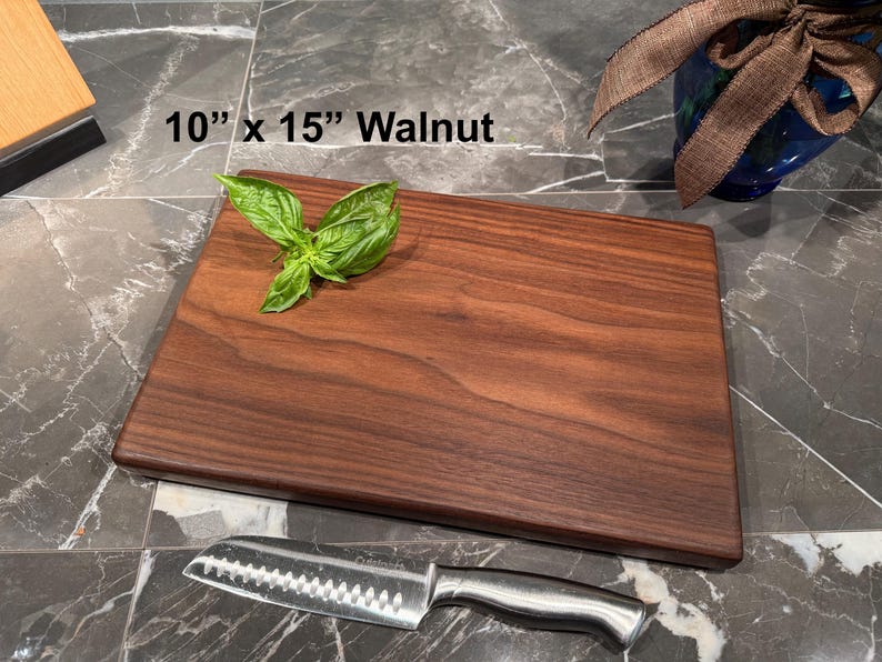May include: A rectangular walnut cutting board measuring 10 inches by 15 inches. The dark brown wood board features a natural grain pattern and is topped with a small sprig of fresh green basil leaves. A stainless steel santoku knife is placed below.