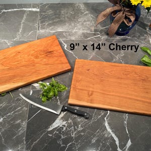 May include: Two rectangular cherry wood cutting boards measuring 9 inches by 14 inches. The boards have a smooth, reddish-brown grain. A stainless steel chef knife with a black handle and fresh green basil leaves are placed near the boards.