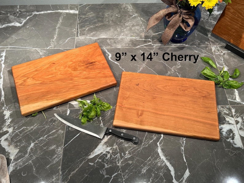 May include: Two rectangular cherry wood cutting boards measuring 9 inches by 14 inches. The boards have a smooth, reddish-brown grain. A stainless steel chef knife with a black handle and fresh green basil leaves are placed near the boards.