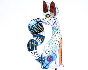 May include: A hand-painted wooden fox figurine with intricate patterns and vibrant colors. The fox features a white base with detailed designs in blue, yellow, and black. Its tail is curled, and it holds a blue and black ball.