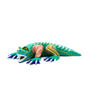 May include: A green, blue, and orange hand-painted wooden lizard figurine with a toothy grin. The lizard is lying on its belly with its legs outstretched.