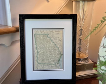 May include: A framed map of Georgia, featuring a black wooden frame and a white mat. The map is in shades of green and beige, with the word "GEORGIA" at the top. The frame is displayed on a dark wooden surface.