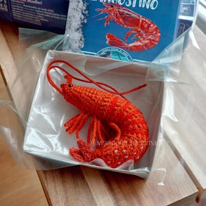 May include: An orange crochet shrimp sits in a white box, with a blue box of frozen shrimp in the background. The box reads "LANGOSITNOS" and "SHRIMPS". A smaller box with the word "LANGOSITNO" and a shrimp illustration is also visible.