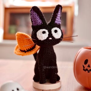 Op de afbeelding: Een handgemaakte zwarte gehaakte kat met paars gevoerde oren en witte ogen, met een oranje heksenhoed. Een kleine pompoen en een spookfiguur zijn ook zichtbaar. De kat zit op een houten voet.