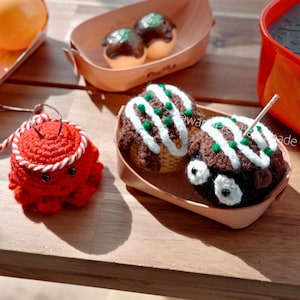 May include: A wooden table displays several handmade crochet items. There are two servings of takoyaki, a red octopus, and a container of orange balls. The takoyaki are brown with white and green accents. The octopus is red with white and red accents.