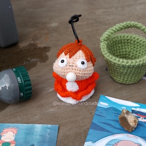 Op de afbeelding: Een gehaakte amigurumi-pop met rood haar en een rood-witte outfit. De pop zit op een tafel met een groene gehaakte mand en een paar geprinte kaarten.