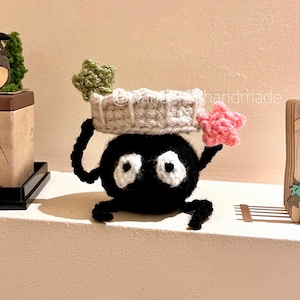 May include: A handmade black crochet character with white eyes and a small mouth. It is wearing a light gray crocheted headband with a green and pink star on top. The character is holding up its arms.