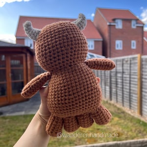 May include: A brown crocheted stuffed animal with grey horns. The animal is being held in a hand with the background out of focus.
