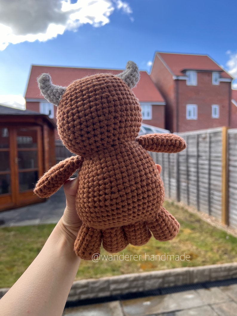 May include: A brown crocheted stuffed animal with grey horns. The animal is being held in a hand with the background out of focus.