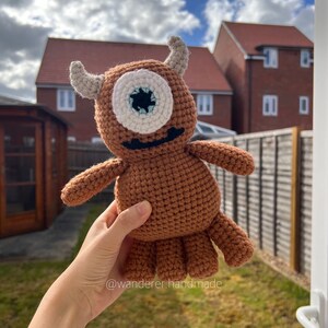 May include: A brown crocheted one-eyed monster with white yarn for the eye and a black mouth. The monster is being held by a hand in front of a green lawn and a house.