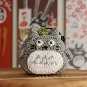 May include: A gray crocheted Totoro plushie with a white belly and a green leaf on its head. The plushie is sitting on a wooden surface.