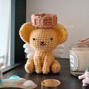 Puede incluir: Una figura de perro de ganchillo hecha a mano con un halo y alas, que lleva una corona marrón. El perro es de color marrón claro con ojos negros y una nariz cosida en negro. El fondo incluye una vela, una pequeña figura de luchador de sumo y un amuleto de madera japonés.