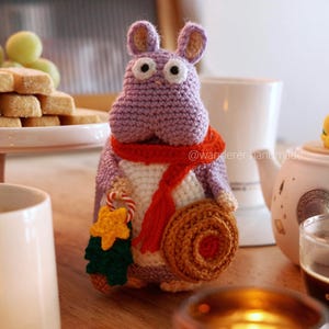Puede incluir: Un juguete de hipopótamo de crochet morado que sostiene un pequeño árbol de Navidad y un bastón de caramelo. El hipopótamo lleva una bufanda roja y un sombrero marrón. El juguete está sentado en una mesa de madera con una taza de café y una tetera en el fondo.