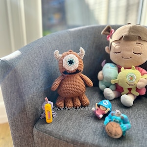 May include: A brown crocheted monster with one large blue eye, a pink crocheted character with a blue hat, a yellow crocheted character with a green eye, and a blue crocheted character with a brown eye. The characters are sitting on a grey armchair. The text "@wanderer_handmade" is visible in the image.