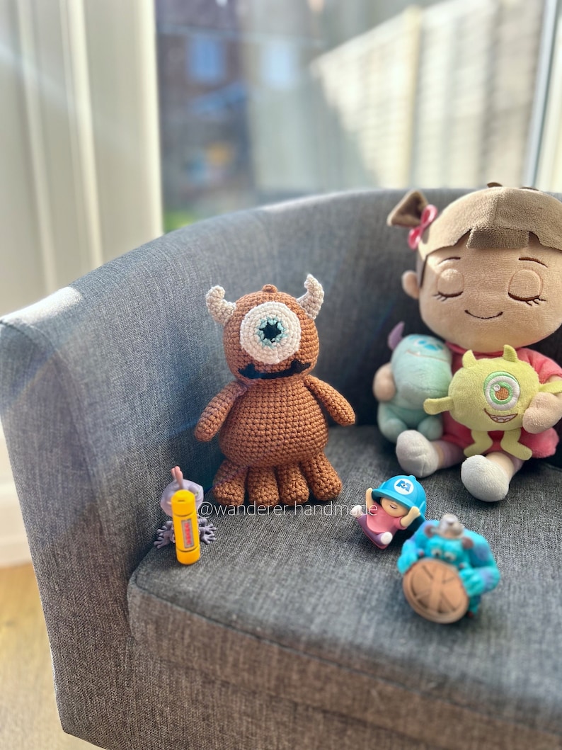 May include: A brown crocheted monster with one large blue eye, a pink crocheted character with a blue hat, a yellow crocheted character with a green eye, and a blue crocheted character with a brown eye. The characters are sitting on a grey armchair. The text "@wanderer_handmade" is visible in the image.