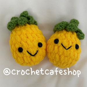 Op de afbeelding: Twee gehaakte ananas knuffels met lachende gezichten. De ananas zijn geel met groene toppen. De knuffels liggen op een witte achtergrond. @crochetcafeshop