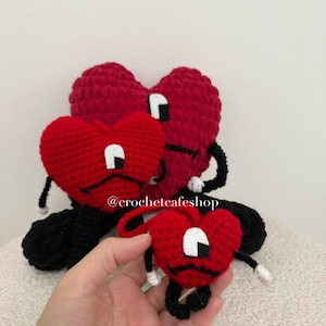 Op de afbeelding: Drie rode gehaakte hartvormige pluche figuren van verschillende groottes, elk met zwarte ledematen, witte handen en een wit en zwart ooglapje-ontwerp. De tekst @crochetcafeshop is zichtbaar over het midden van het grootste hartfiguur.
