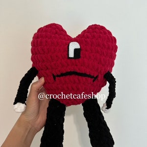 Op de afbeelding: Een rood gehaakt pluche speelgoed in de vorm van een hart met zwarte armen en benen. Het speelgoed heeft een wit en zwart oog en een zwarte frons. Het speelgoed wordt vastgehouden door een hand. De tekst '@crochetcafeshop' is zichtbaar in de afbeelding.