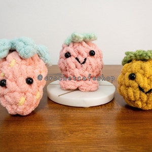 May include: Three crocheted plush toys shaped like a strawberry, pineapple, and a generic fruit with a green leaf on top. The strawberry is pink with a light blue leaf, the pineapple is yellow with a green top, and the generic fruit is pink with a green leaf. All three toys have black eyes and a smiling mouth.