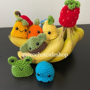 May include: A collection of crocheted fruit and animal plushies, including a strawberry, pear, orange, avocado, frog, and a blue blob, displayed on a bunch of bananas. The plushies have embroidered faces and are made with colorful yarn. The image includes the text "@crochetcafeshop".