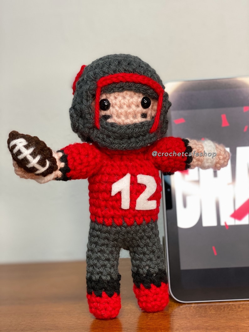 K&ouml;nnte beinhalten: Geh&auml;kelte Footballspieler-Puppe. Die Puppe tr&auml;gt ein rotes Trikot mit der Nummer 12, eine graue Hose und einen grauen Helm mit roten Akzenten. Sie h&auml;lt einen braunen und wei&szlig;en Football. Das Gesicht der Puppe ist hellrosa mit schwarzen Augen und schwarzen Augenstreifen.