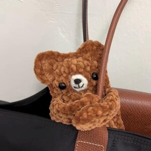 May include: A brown crocheted teddy bear peeking out of a black tote bag with brown leather handles.