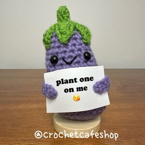 May include: A purple crocheted eggplant holding a white sign that says "plant one on me" with a blushing emoji. The eggplant is standing on a wooden base.