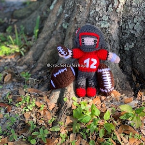 K&ouml;nnte beinhalten: Handgemachte Footballspieler-Puppe aus H&auml;kelarbeit, die ein rotes Trikot mit der Nummer 12, einen grauen Helm tr&auml;gt und einen braunen Football h&auml;lt. Die Puppe steht in der N&auml;he eines Baumstamms, mit zwei weiteren Fu&szlig;b&auml;llen in der N&auml;he. Die Puppe ist etwa 15 cm gro&szlig;.