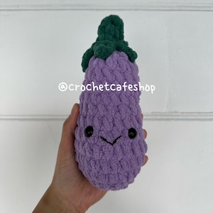 Op de afbeelding: Een paarse gehaakte aubergine met een groene gehaakte stengel en een glimlachend gezicht. De aubergine wordt in een hand gehouden. De tekst "@crochetcafeshop" is zichtbaar in de afbeelding.