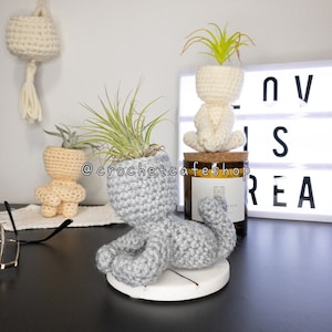 May include: Three crocheted planter pots in the shape of people, each holding a small plant. The pots are in shades of gray and beige. The background features a light box with the text "LOVE IS REAL" and a smiley face.