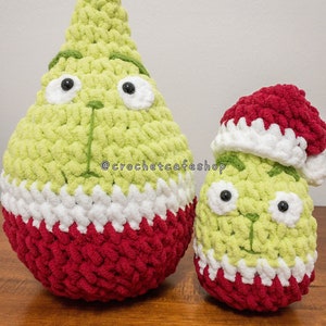 May include: Two crocheted green and red Christmas ornaments with white accents. The larger ornament has a pointed top and the smaller ornament has a red crocheted hat. The ornaments have black button eyes and a crocheted smile.