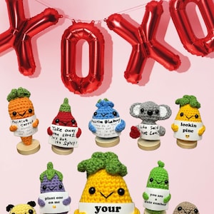 May include: A collection of crocheted amigurumi vegetables holding signs with Valentine's Day messages. The vegetables include a carrot, a chili pepper, a blueberry, a koala, a pineapple, an eggplant, a yellow pepper, a cucumber, and a potato. The signs say "Positive Carrot", "Take away the stress, it's Hot, it's Spicy!", "Positive Blueberry", "Who Said Love Love", "Lookin Pine", "Plant one on me", "Your text here", "You are a cute cumber", and "You are one hot potato".