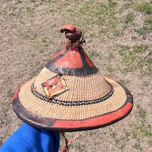 May include: A large, conical straw hat with a leather brim and a red, black and brown design. The hat has a small, round leather top with a decorative design.