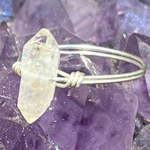May include: A clear quartz crystal wrapped in silver wire, set on a ring band. The ring is displayed on a bed of purple amethyst crystals.