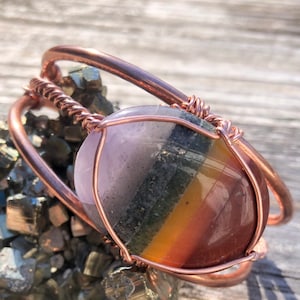 May include: A copper wire wrapped bracelet with a multi-colored gemstone. The gemstone has shades of purple, grey, yellow, orange, and red. The bracelet is sitting on a bed of pyrite crystals.