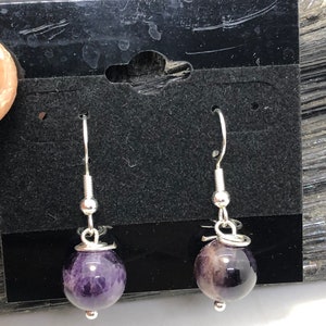May include: A pair of silver earrings with purple amethyst stone beads. The earrings are on a black card with a hole for hanging.