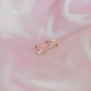 May include: A pair of small, gold-colored cross stud earrings. The earrings are displayed on a pink, satin-like fabric. The crosses are simple in design and appear to be made of metal.