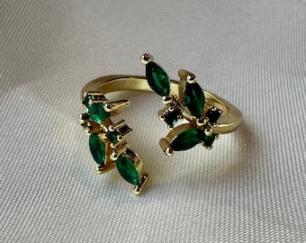 May include: A gold ring with a split design featuring emerald green gemstones. The ring is open and adjustable.