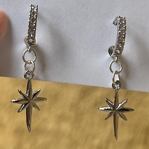 Puede incluir: Un par de pendientes de aro plateados con colgantes de estrella. Los aros están incrustados con pequeños cristales transparentes. Los pendientes se muestran en una tarjeta blanca con el texto "23 NORTH" y una dirección web.