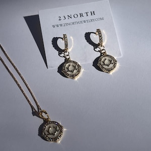 Puede incluir: Collar y pendientes de oro con un colgante blanco que presenta una silueta de ciervo. El colgante y los pendientes están rodeados de pequeños diamantes de imitación transparentes. La joyería se muestra en una tarjeta blanca con el texto "23NORTH www.23northjewelry.com".